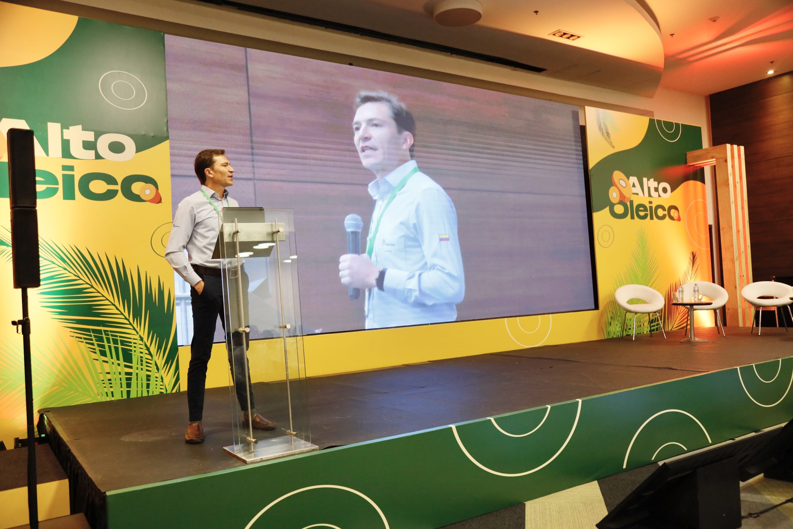 Hombre de pie en un podio en un escenario, con una gran pantalla LED de fondo que muestra un primer plano de él mismo hablando.