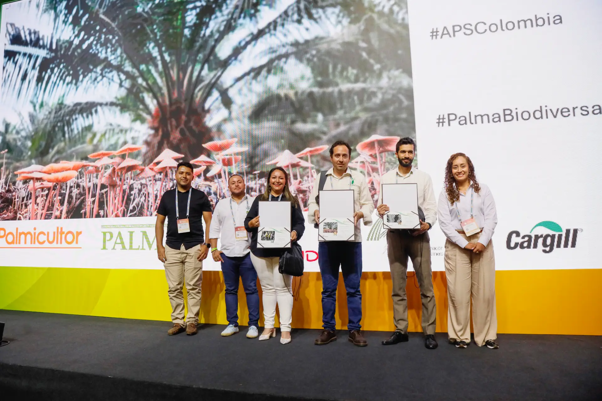 Personas con certificados, de pie en escenario que muestra los hashtags #APSColombia y #PalmaBiodiversa.