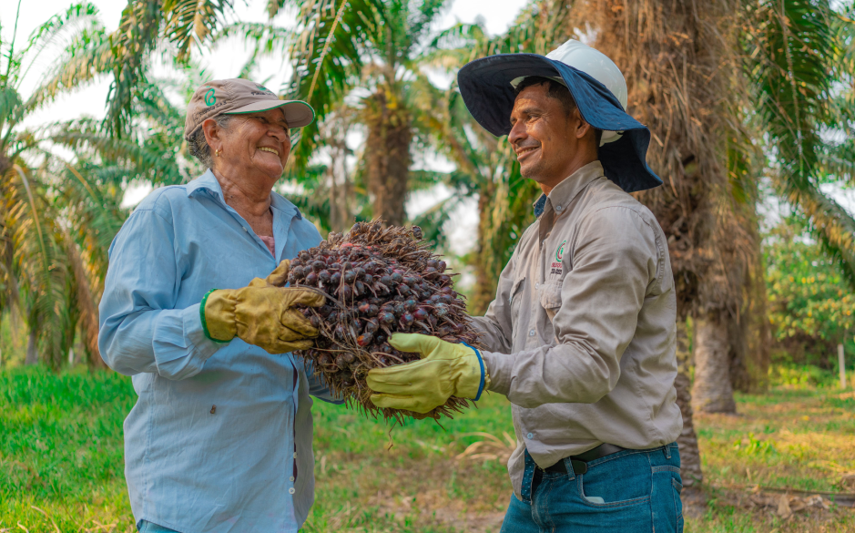 Dos agricultores sonrientes sostienen un racimo de frutos de palma de aceite en una plantación.