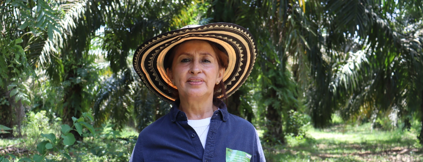 Retrato de una mujer con un sombrero vueltiao tradicional, parada en medio de una plantación de palma de aceite bajo el sol.