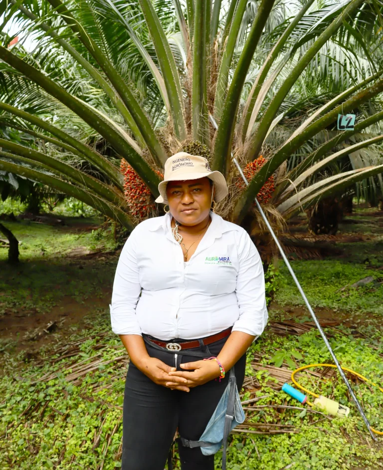 Mujer con camisa blanca y sombrero, de pie frente a una palma de aceite con frutos.