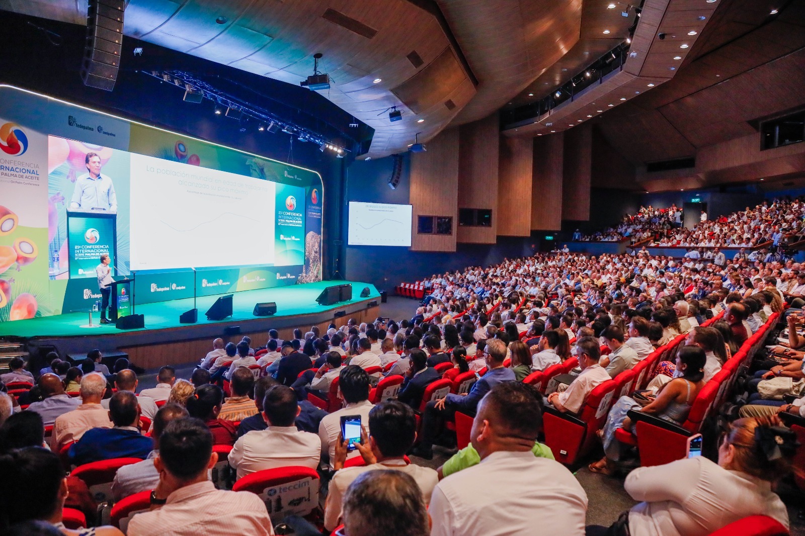vista panorámica de un gran auditorio lleno de gente, con un orador en el escenario presentando frente a una gran pantalla.